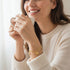A smiling woman in a cream sweater holds a white mug, wearing a 14k gold-filled bracelet with protective elephant and Figa hand charms.